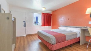 Quarto de hotel com cama, escrivaninha, cadeira, janela com cortinas e porta.