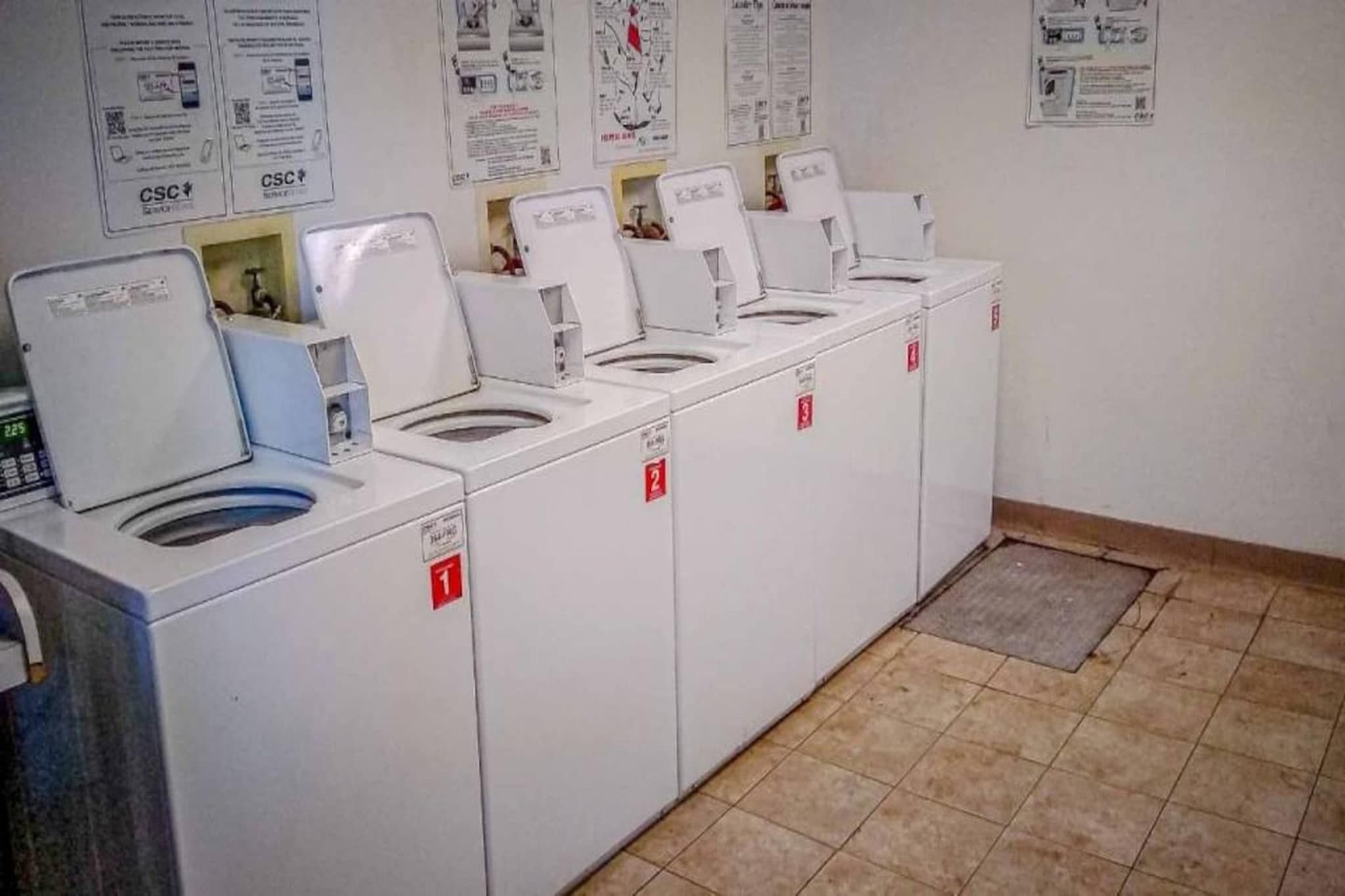 A row of white laundry machines with numbered doors, a tiled floor, and informational posters on the wall.