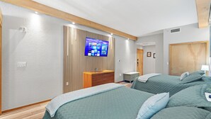 Quarto com duas camas, televisão fixada em uma parede de painéis de madeira, mesa de cabeceira e guarda-roupa.