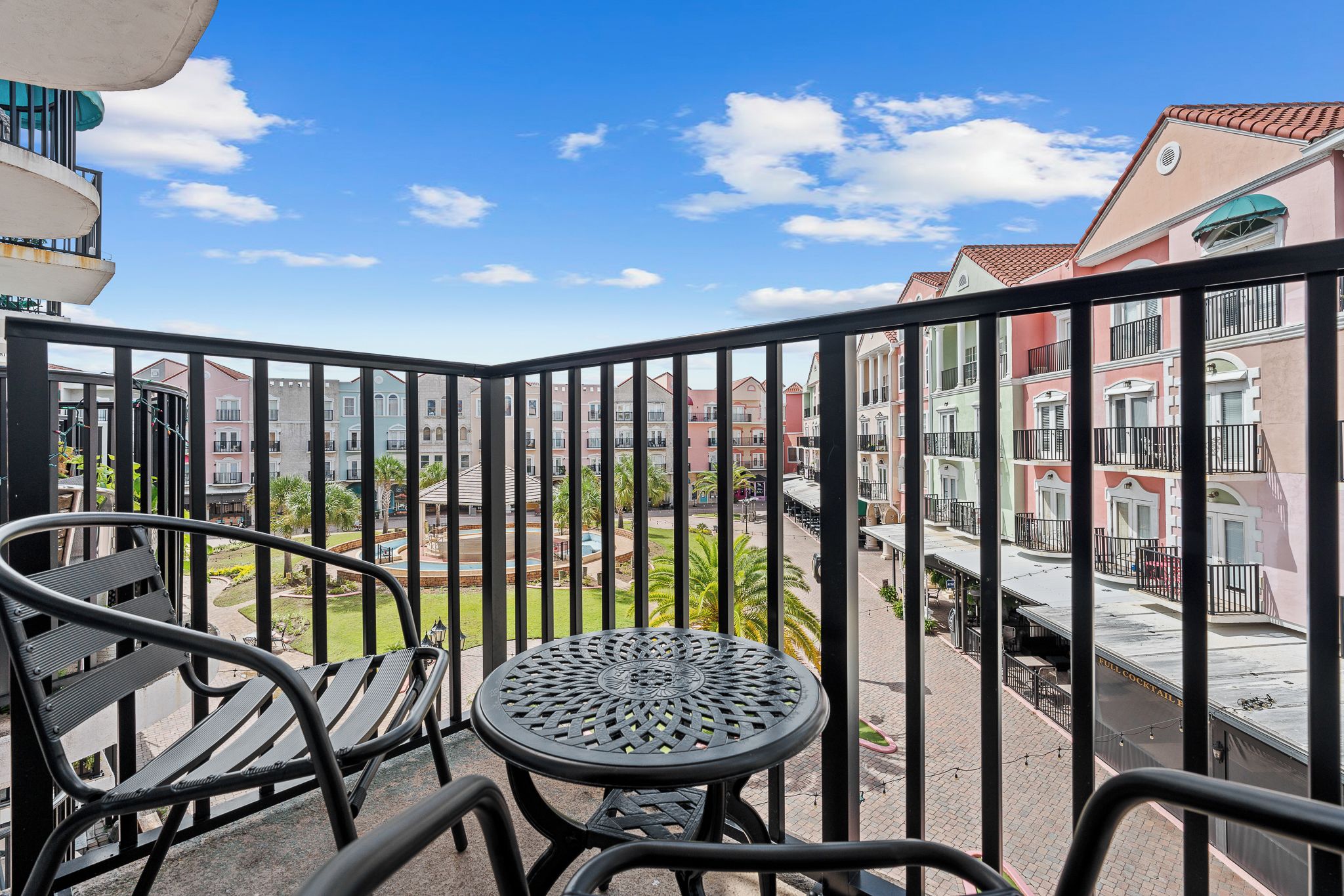 Un balcón con mesa y sillas, con vista a un patio con edificios y un cielo despejado.