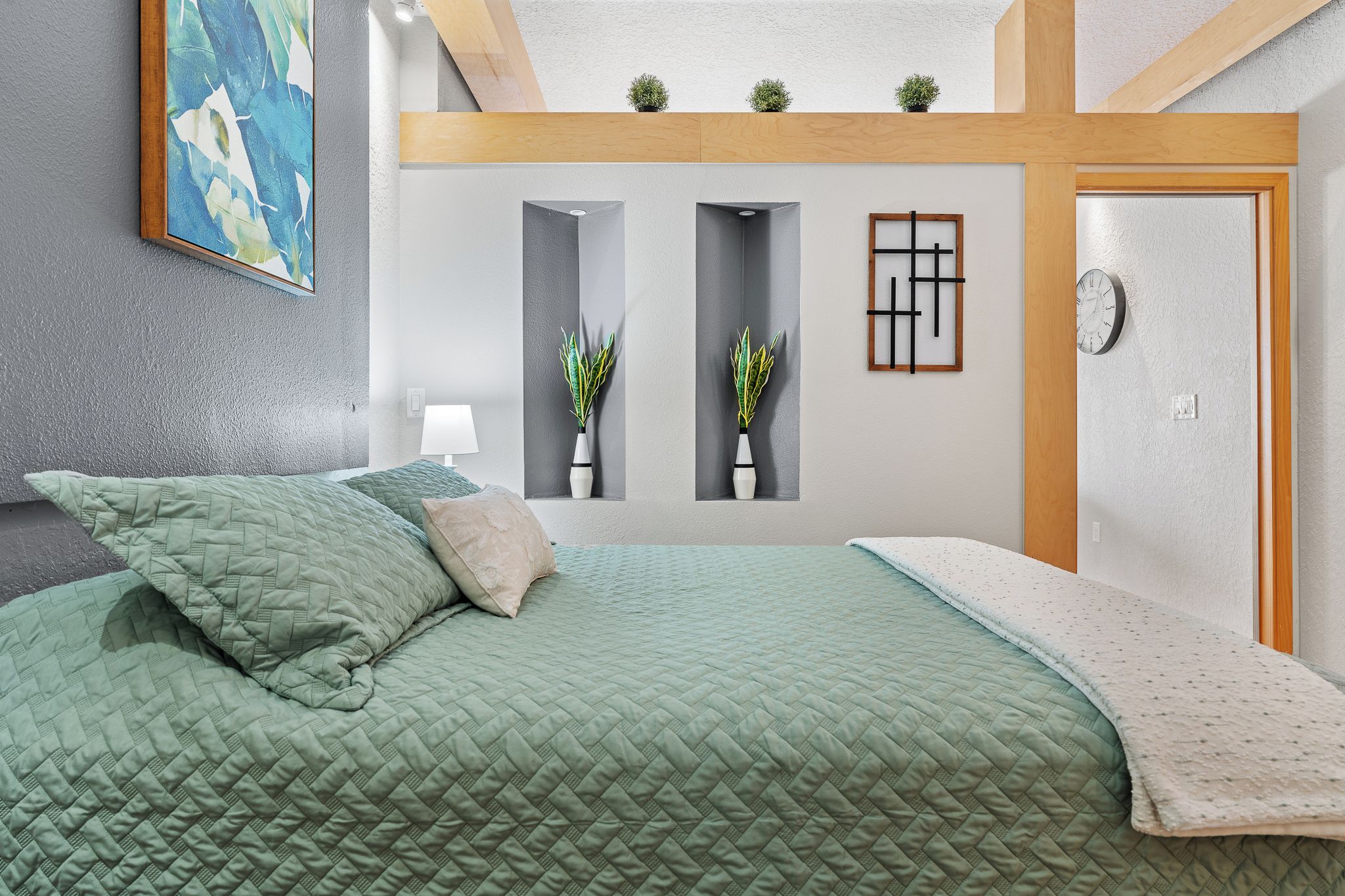 A bedroom with a green quilted bedspread, a bedside lamp, and two potted plants.