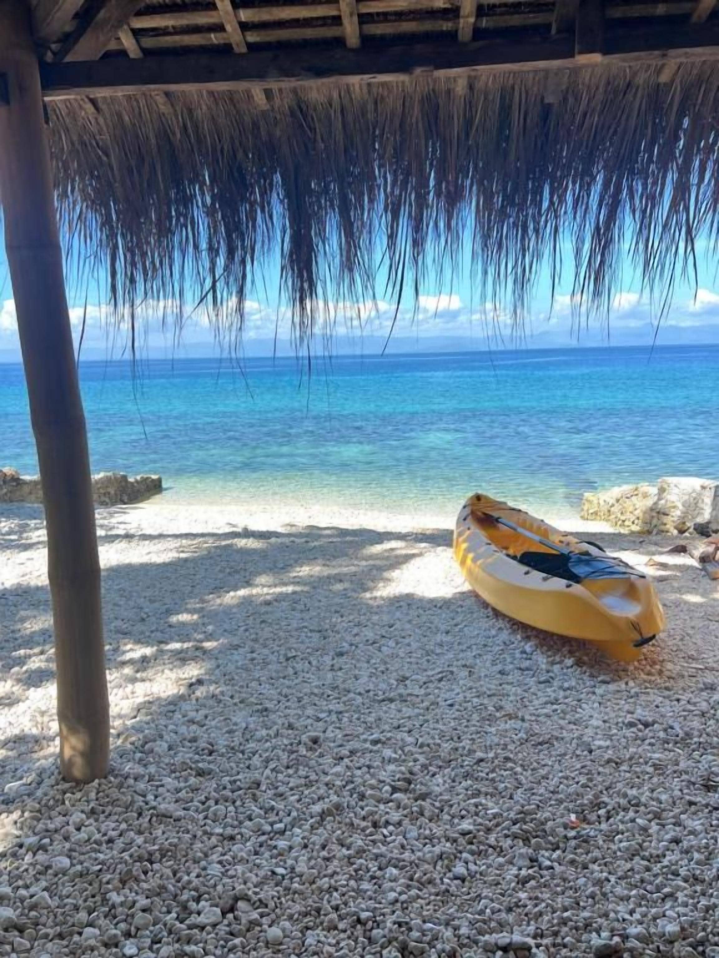 Una playa con una estructura de techo de paja, un kayak amarillo y una orilla pedregosa.