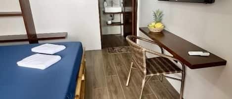 Habitación compacta con cama, escritorio, silla, televisor y baño pequeño.