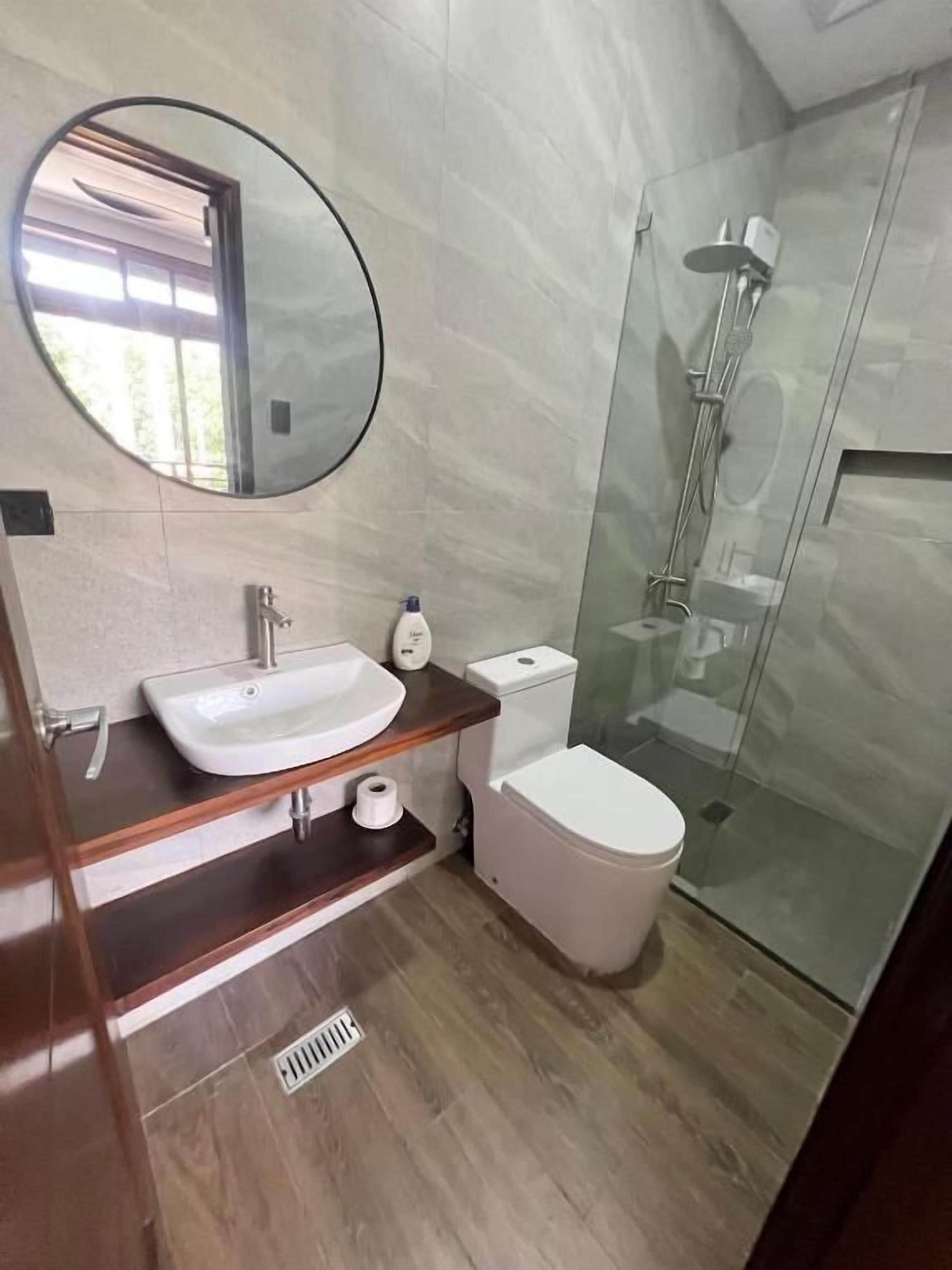 Un baño moderno con un espejo redondo, un lavabo, un inodoro y una ducha con mampara de vidrio.