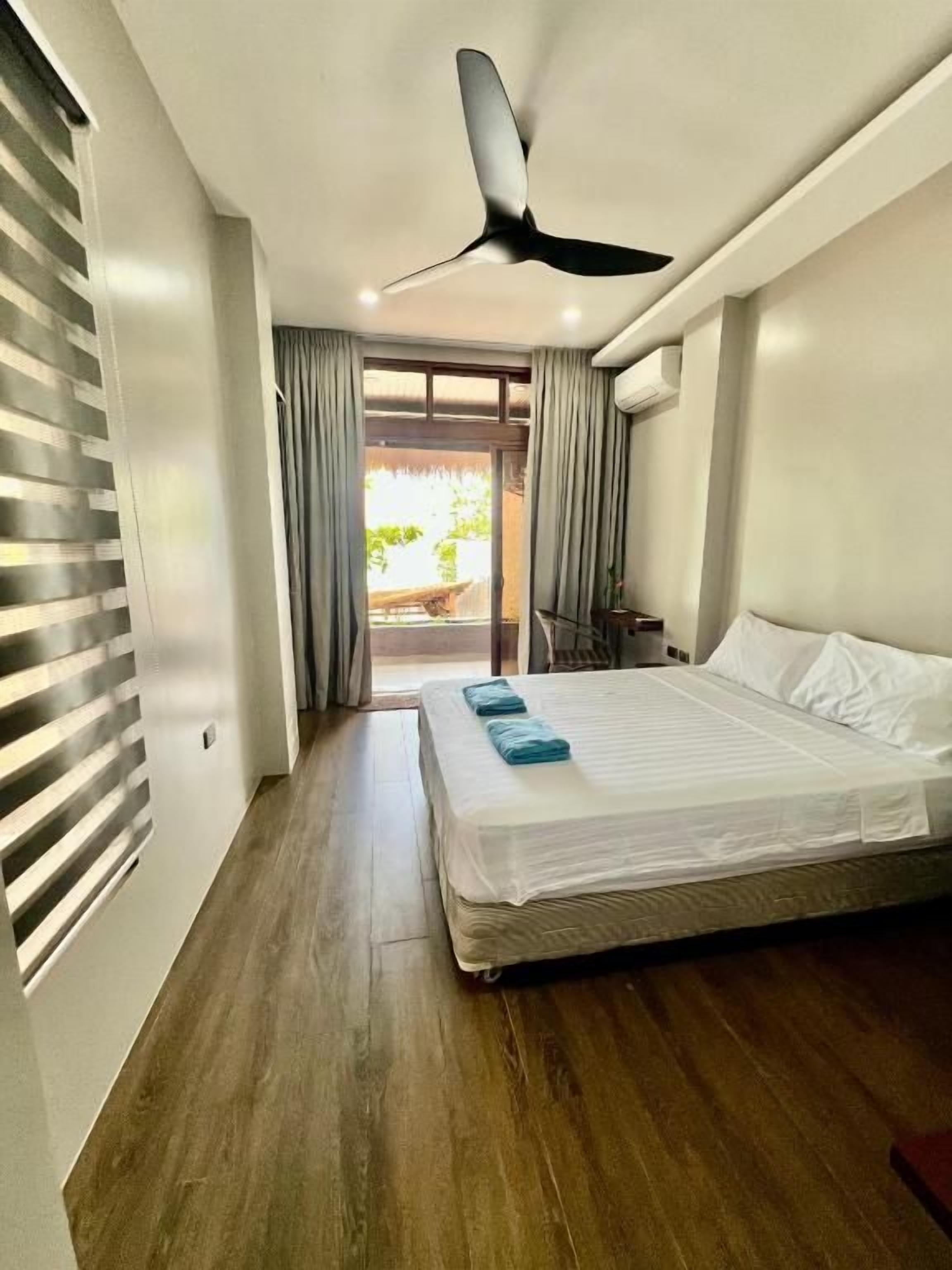 A bedroom with a bed, a ceiling fan, a sliding glass door leading to an outdoor area, and a wooden floor.