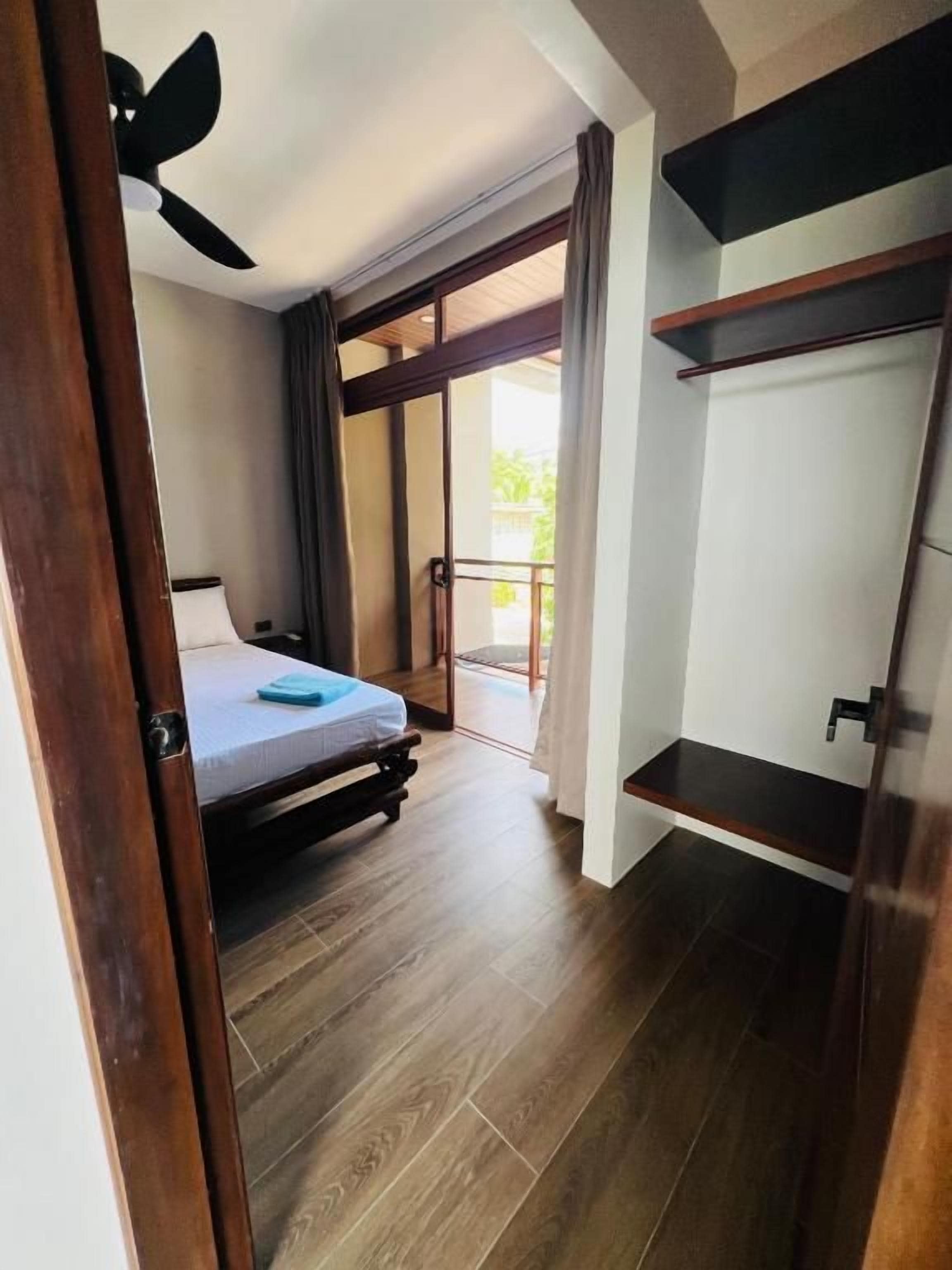 Un dormitorio con una cama, ventilador de techo, puerta corrediza de vidrio que da a un balcón y suelo de madera.
