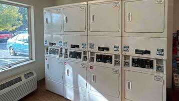 Une buanderie équipée de plusieurs machines à laver et de plusieurs séchoirs, une fenêtre donnant sur les voitures garées et une bouche d’aération dans le mur.