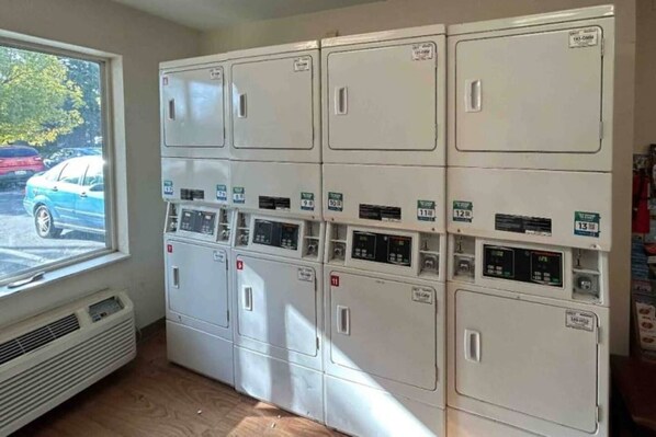 A laundry room with multiple washing machines and dryers, a window with a view of parked cars, and a vent on the wall.