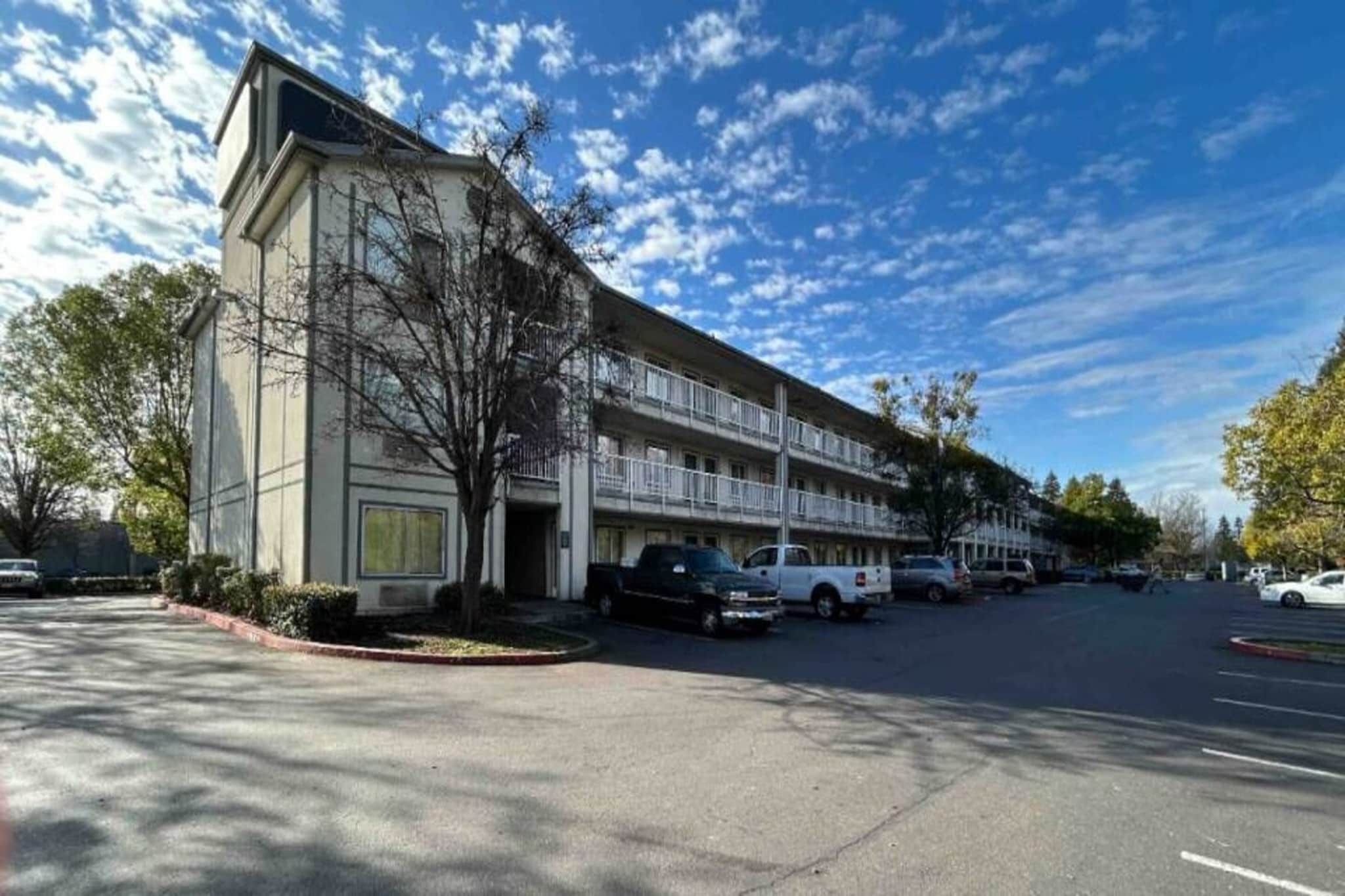 Motel de diversos pisos amb un aparcament, cel blau i arbres.