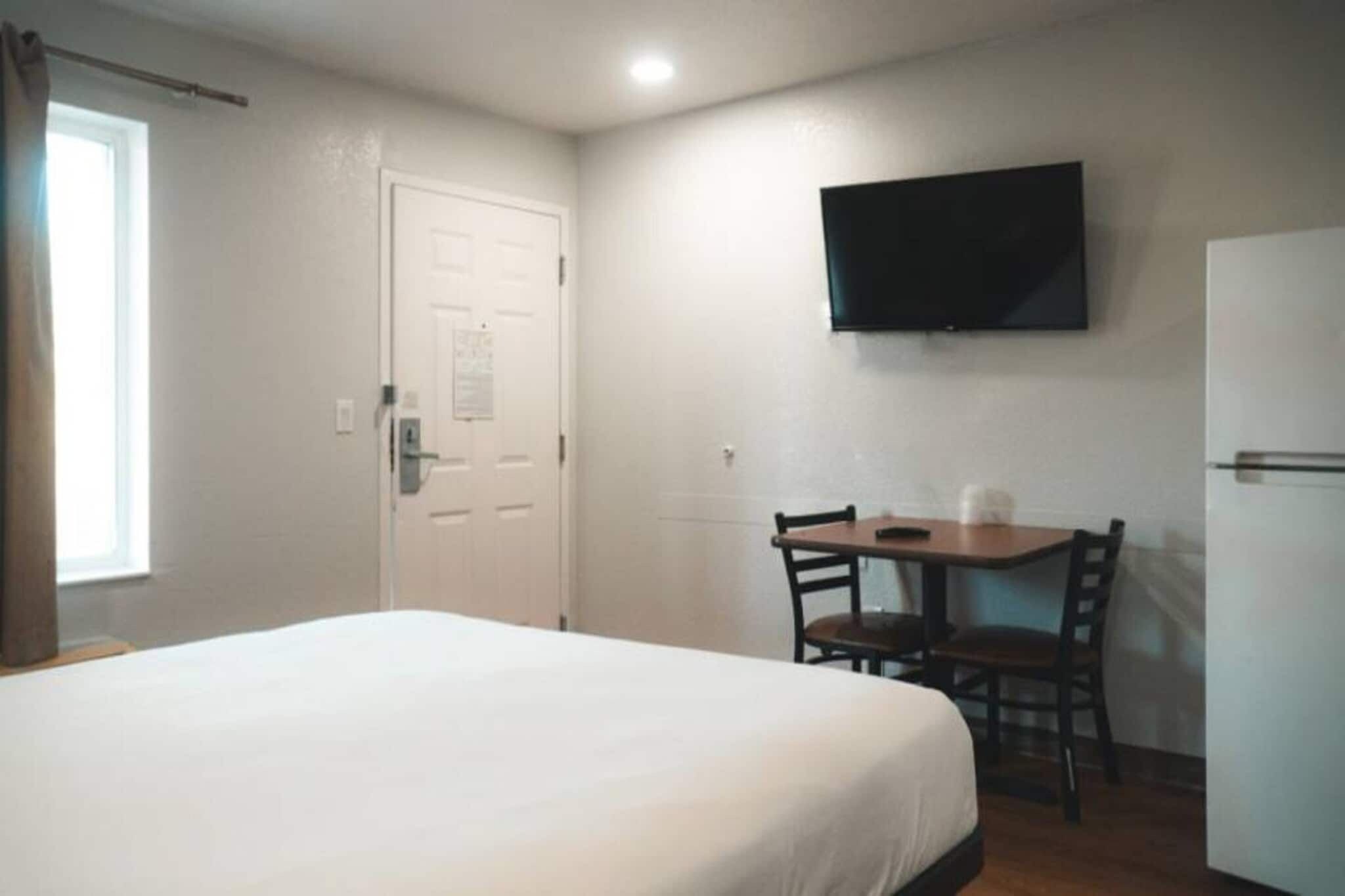 Una habitación de hotel con cama, escritorio con silla, televisión en la pared y una puerta.