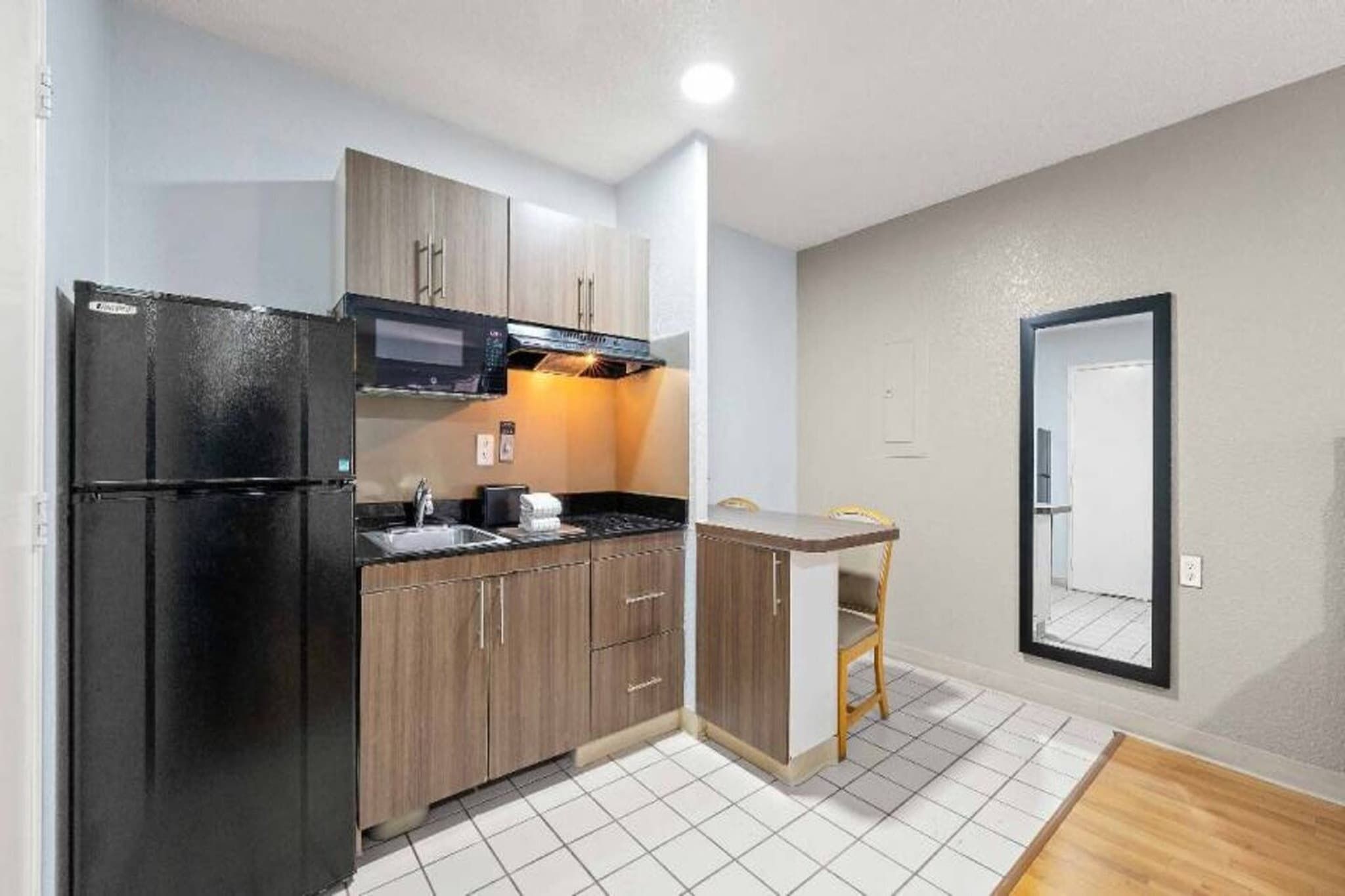 A compact kitchen with a refrigerator, microwave, and stove. There is a small dining area with a chair and a table. The room has white tiled flooring and light-colored walls.
