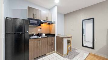 A compact kitchen with a refrigerator, microwave, and stove. There is a small dining area with a chair and a table. The room has white tiled flooring and light-colored walls.