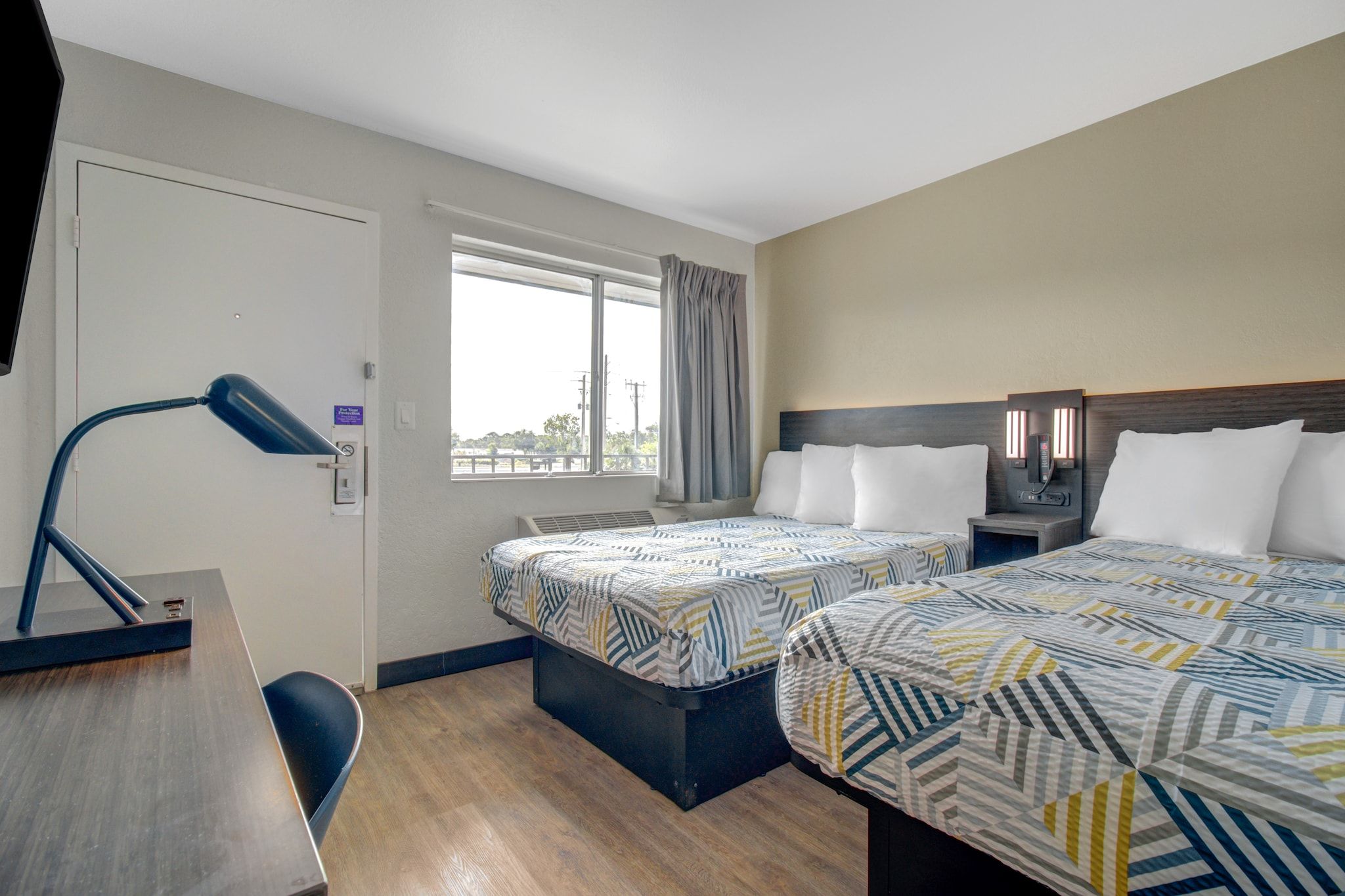 Een hotelkamer met twee bedden, een bureau, een stoel, een raam met gordijnen en een lamp.