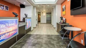 A hotel lobby with a reception desk, a TV screen, a vending machine, a seating area with a table and chairs, and an elevator in the background. - Spacious 1 Queen Bed Non-Smoking (Fort Mill)