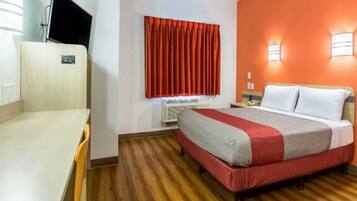 Habitación de hotel con cama, escritorio, televisor, ventana con cortinas y lámpara de pared.