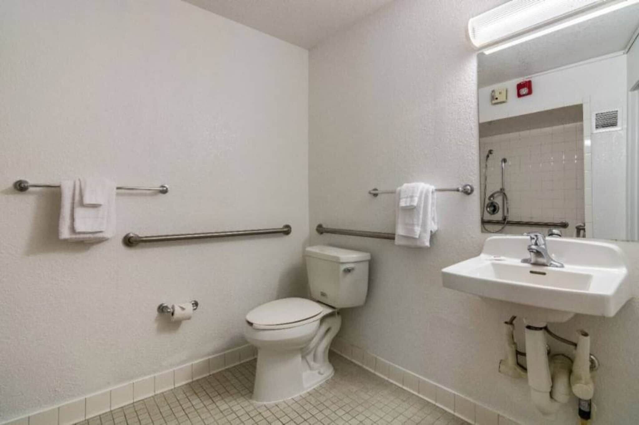 A standard hotel bathroom with a toilet, handrails, a sink, and a mirror.