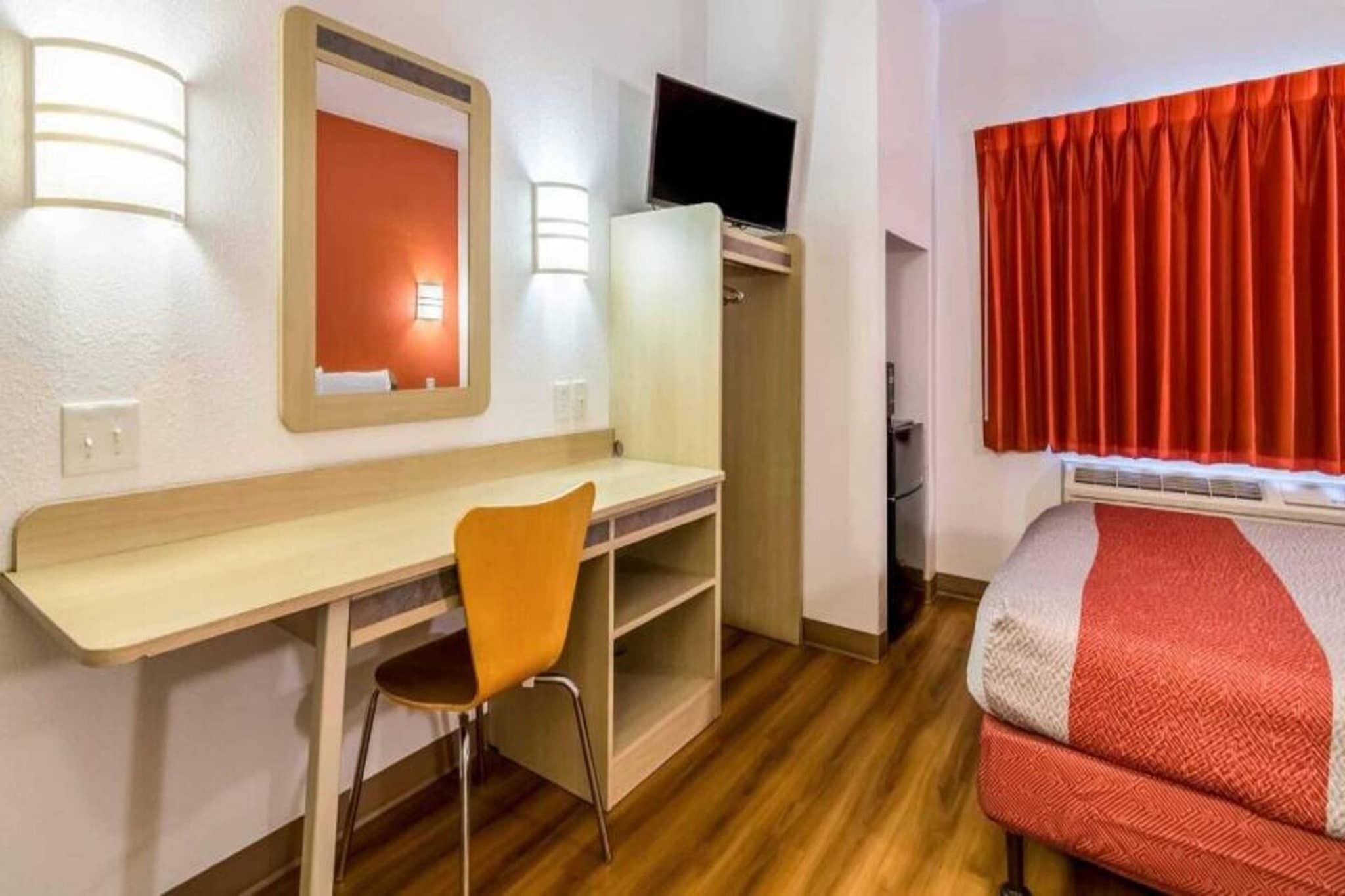 A hotel room with a desk, chair, TV, wardrobe, and a bed with a red and white blanket.