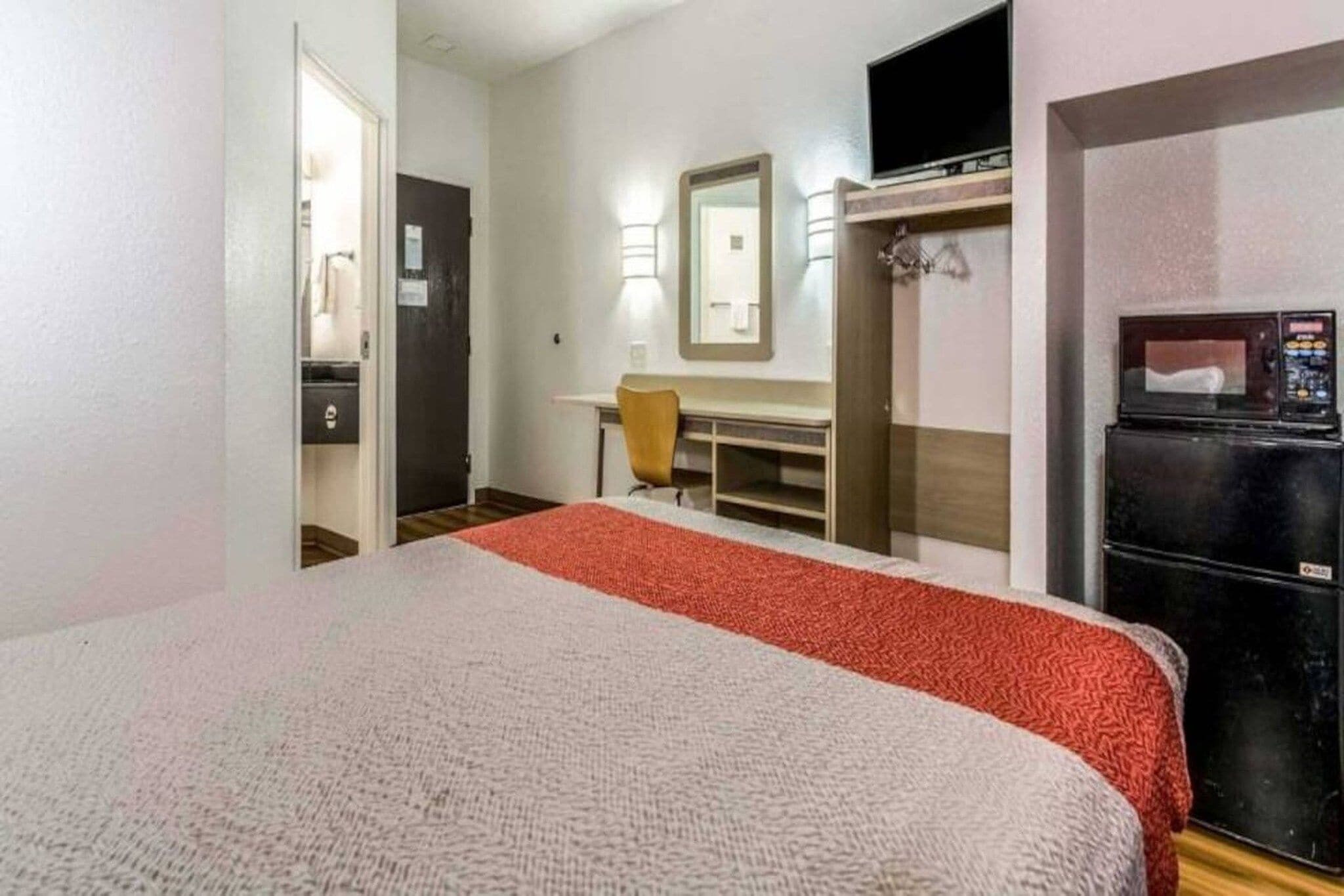 A hotel room with a bed, a desk, a chair, a mirror, a wardrobe, and a refrigerator.