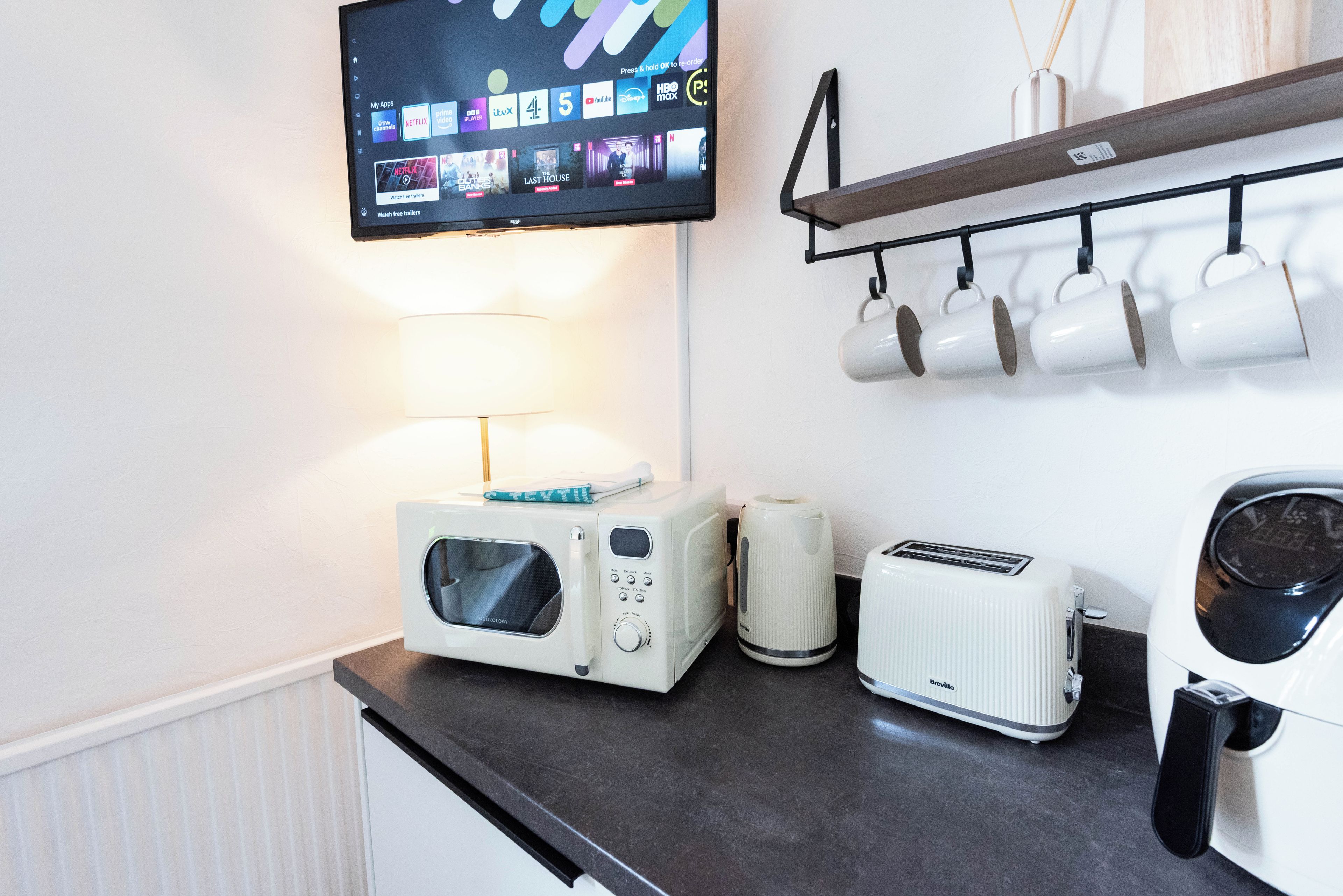 A kitchenette with a microwave, toaster, and kettle on a countertop, a TV mounted above, and a shelf with mugs.