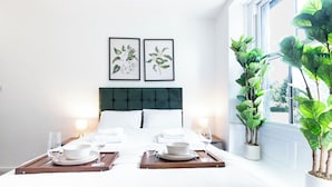 Quarto de hotel com cama bem arrumada, duas gravuras de plantas emolduradas na parede, uma planta grande em vaso junto à janela e uma bandeja com copos e tigelas.