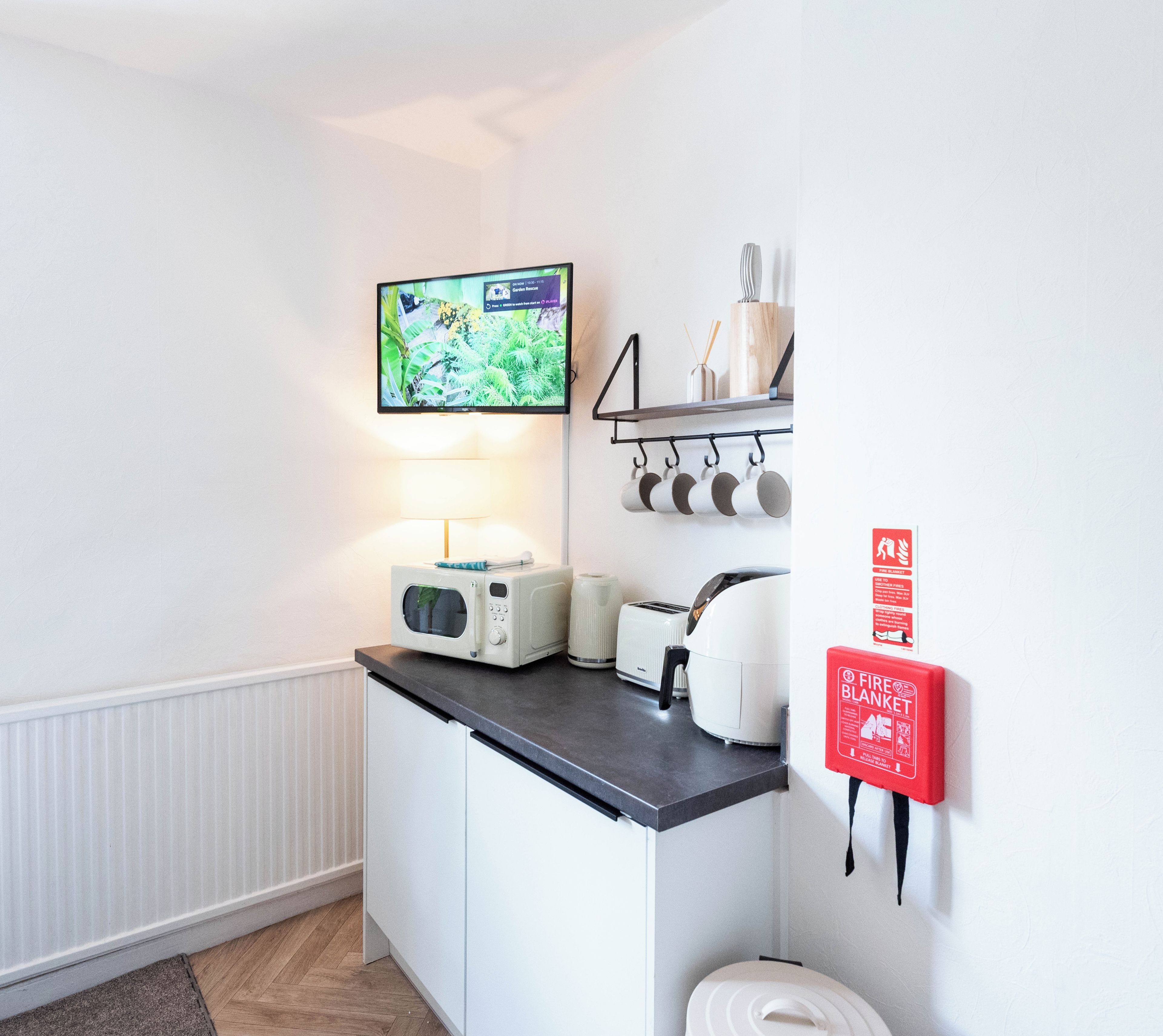 A compact kitchenette with a microwave, toaster, and kettle on the counter, a small shelf with cups, a fire blanket, and a fire extinguisher.