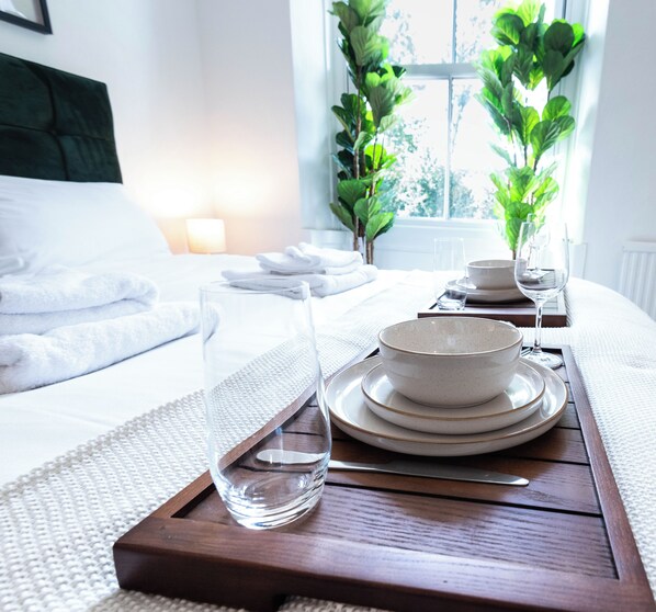 A neatly made bed with white linens, a tray with glassware and bowls, and a window with a view of greenery.