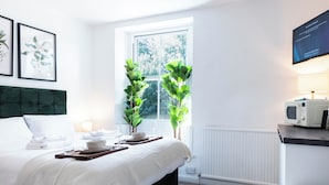 Cama bem arrumada com edredom branco, mesa de cabeceira com abajur e janela com vista para área verde.