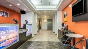 Recepció d'hotel amb taulell d'informació, televisió penjada a la paret, màquina expenedora i zona de seients amb cadires i taula.