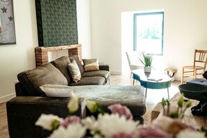A living room with a brick fireplace, a sofa, a coffee table, and a chair. There is a window with a view of trees.