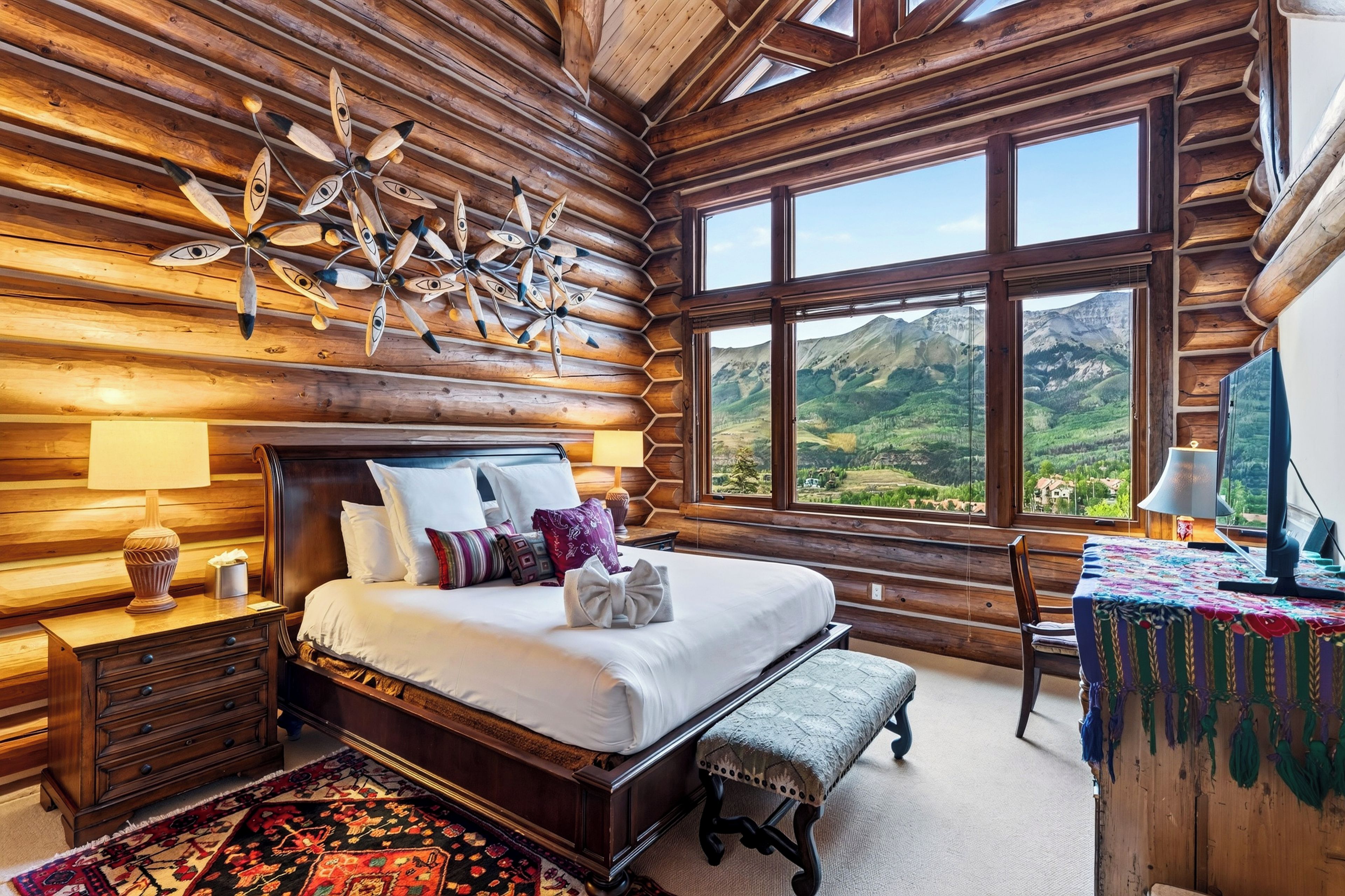 A log cabin-style bedroom with a large bed, wooden headboard, and a view of mountains through large windows.