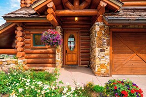 A log cabin with a stone foundation, a wooden front door, and a hanging flower basket. - Log Home • Hot Tub + Full Gym | Ski-in/out Funicular | 115 Tristant (Mountain Village)