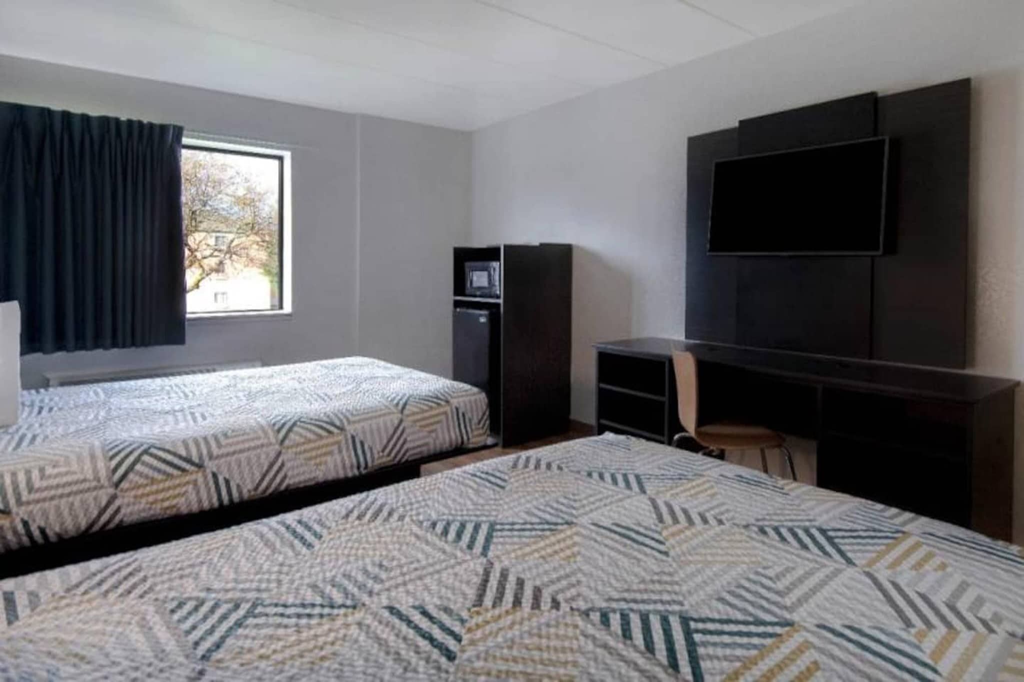A hotel room with two beds, a TV, a small kitchenette, and a window with curtains.