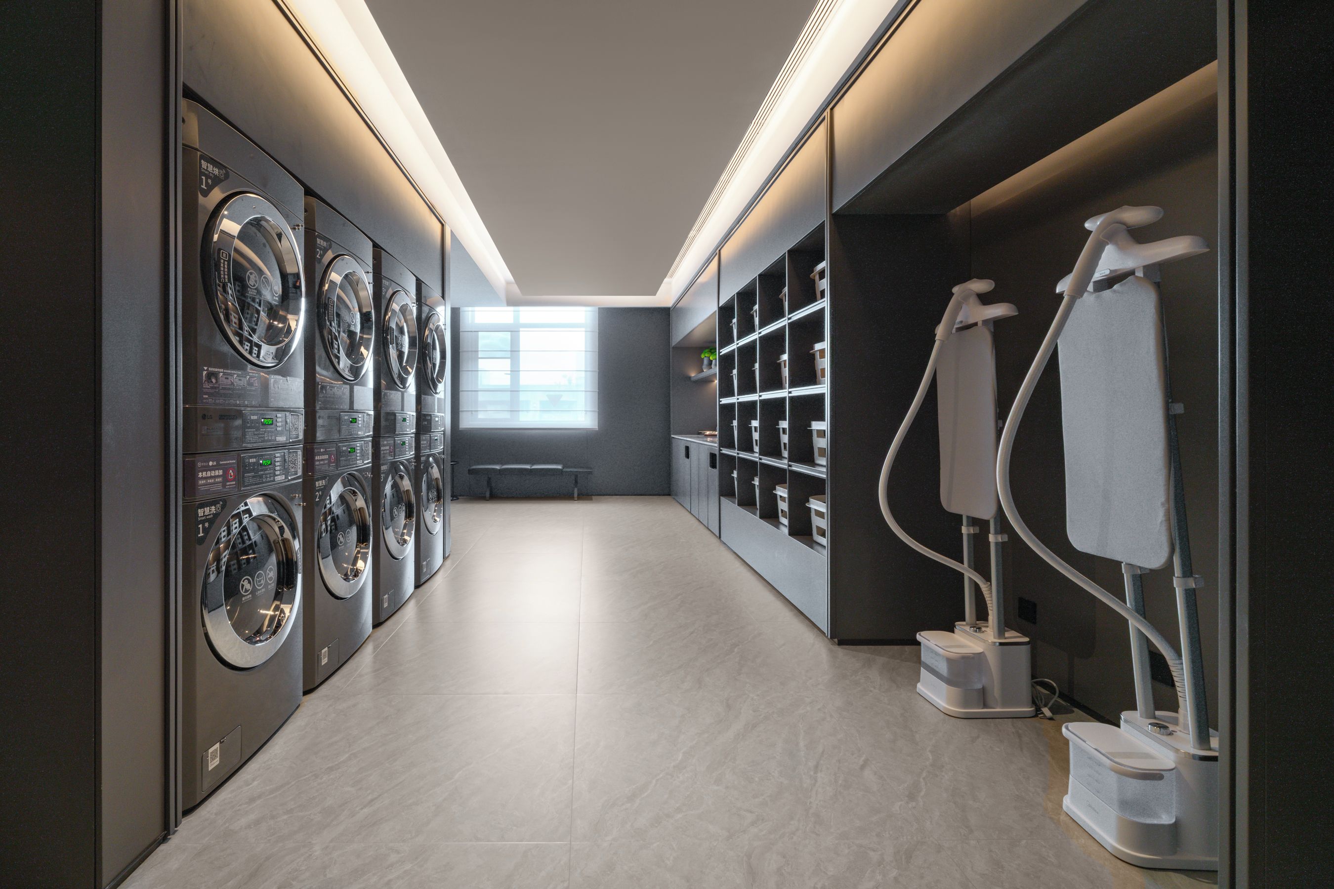 A laundry facility with multiple washing machines and dryers, a bench, and a window providing natural light.