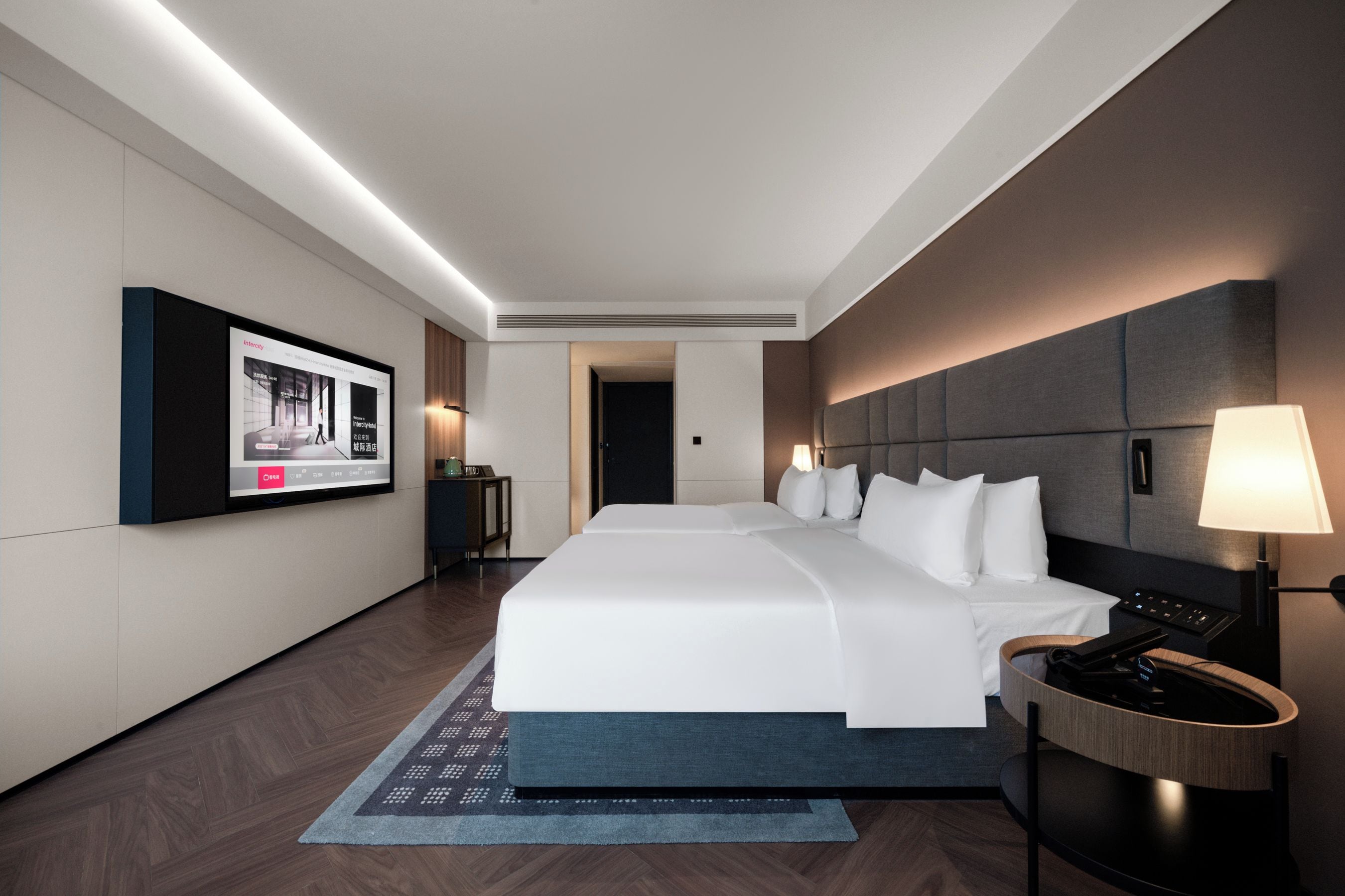 A modern hotel room with a large bed, a flat-screen TV mounted on the wall, a bedside table with a lamp, and a wooden headboard.