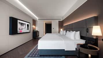 Quarto de hotel moderno com uma cama grande, televisão de tela plana na parede, mesa de cabeceira com luminária e cabeceira de madeira.