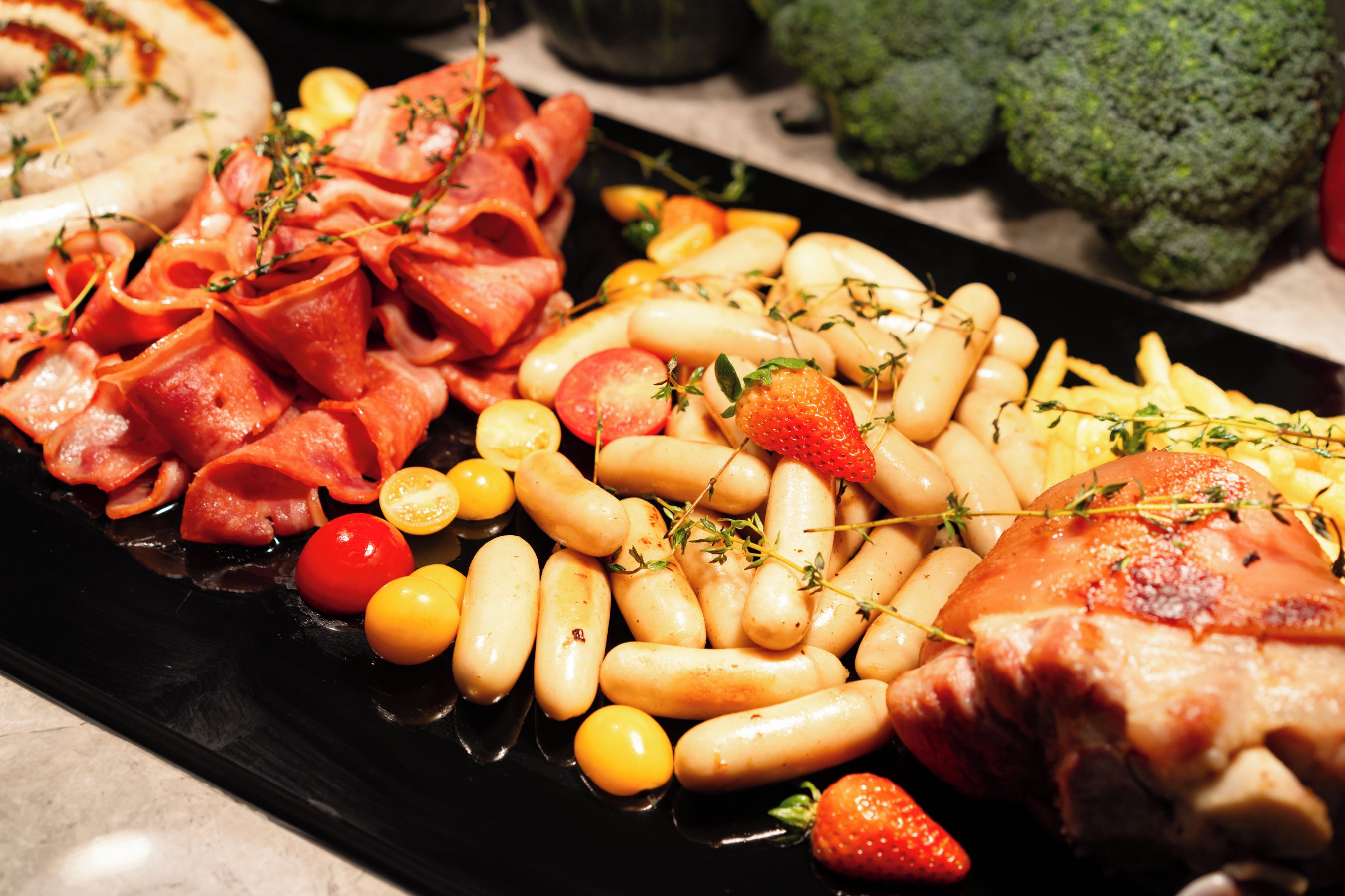 A variety of food items including sausages, bacon, tomatoes, and cheese on a black plate.