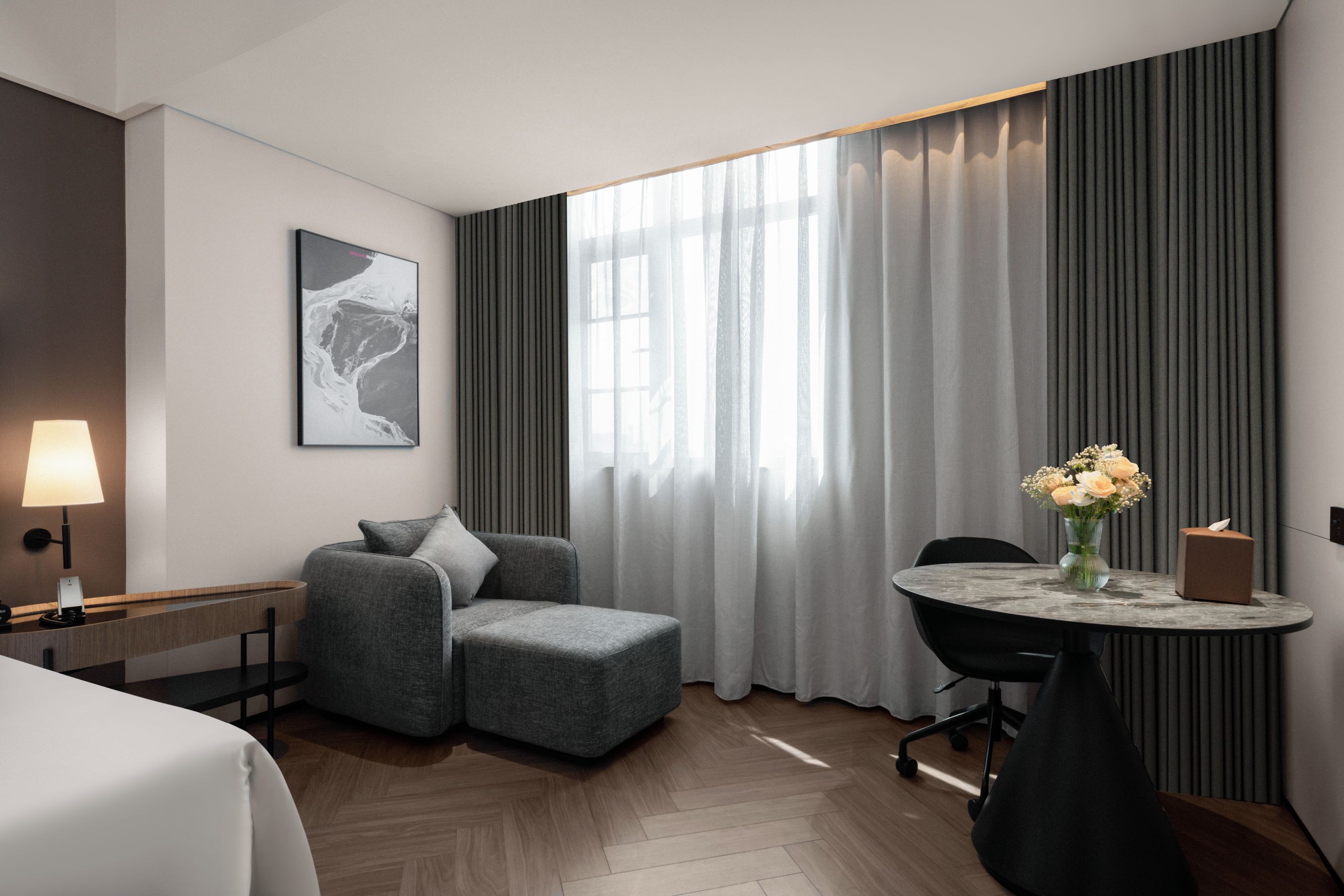 Ein modernes Hotelzimmer mit einem grauen Sofa, einem kleinen runden Tisch mit Blumen, einem Schreibtisch mit einer Lampe und einem großen Fenster mit transparenten Vorhängen.