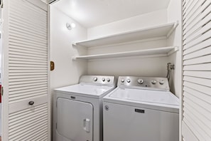 A laundry room with two white wall-mounted washers and dryers, a small shelf above the dryers, and a door with horizontal blinds. - Forest Views + Pool + Pickleball Access | Near Lake + Ski | Tahoe Pines Retreat (Kings Beach)