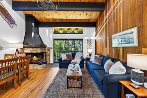 A cozy living room with a stone fireplace, wooden ceiling beams, a blue sofa, and a view of the outdoors. - Forest Views + Pool + Pickleball Access | Near Lake + Ski | Tahoe Pines Retreat (Kings Beach)