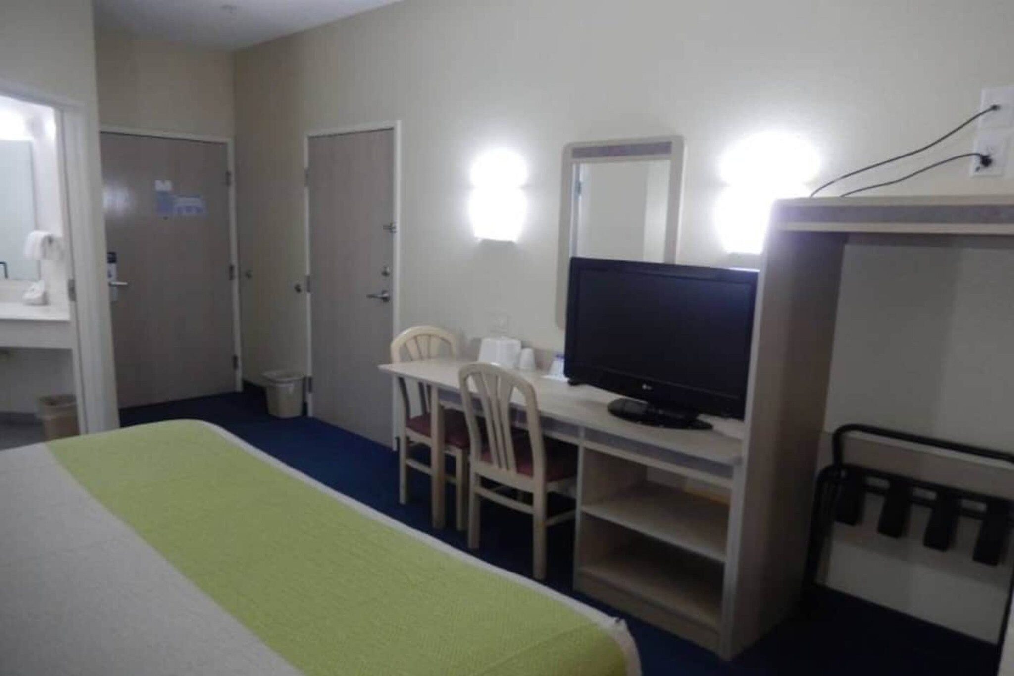 A hotel room with a bed, a desk with a computer, a chair, and a TV. There are two doors, one leading to a bathroom and the other to a closet.
