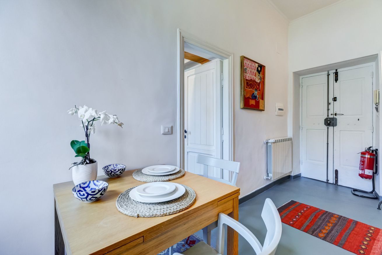 A dining area with a wooden table set for two, white chairs, a vase with white flowers, and a painting on the wall.