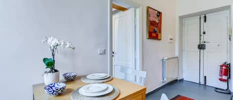 A dining area with a wooden table set for two, white chairs, a vase with white flowers, and a painting on the wall.
