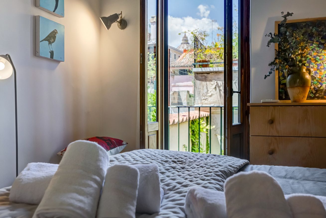 A bedroom with a bed, a nightstand, a lamp, a dresser, and a window with a view of buildings and greenery.