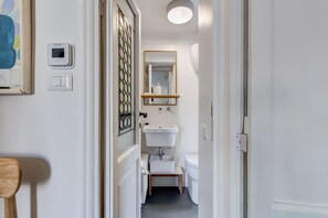 Ein schmaler Flur führt zu einem Badezimmer mit einem weißen Waschbecken, einer Toilette und einer kleinen Ablagetreppe. Über dem Waschbecken hängt ein Spiegel und an der Wand befindet sich ein Thermostat.