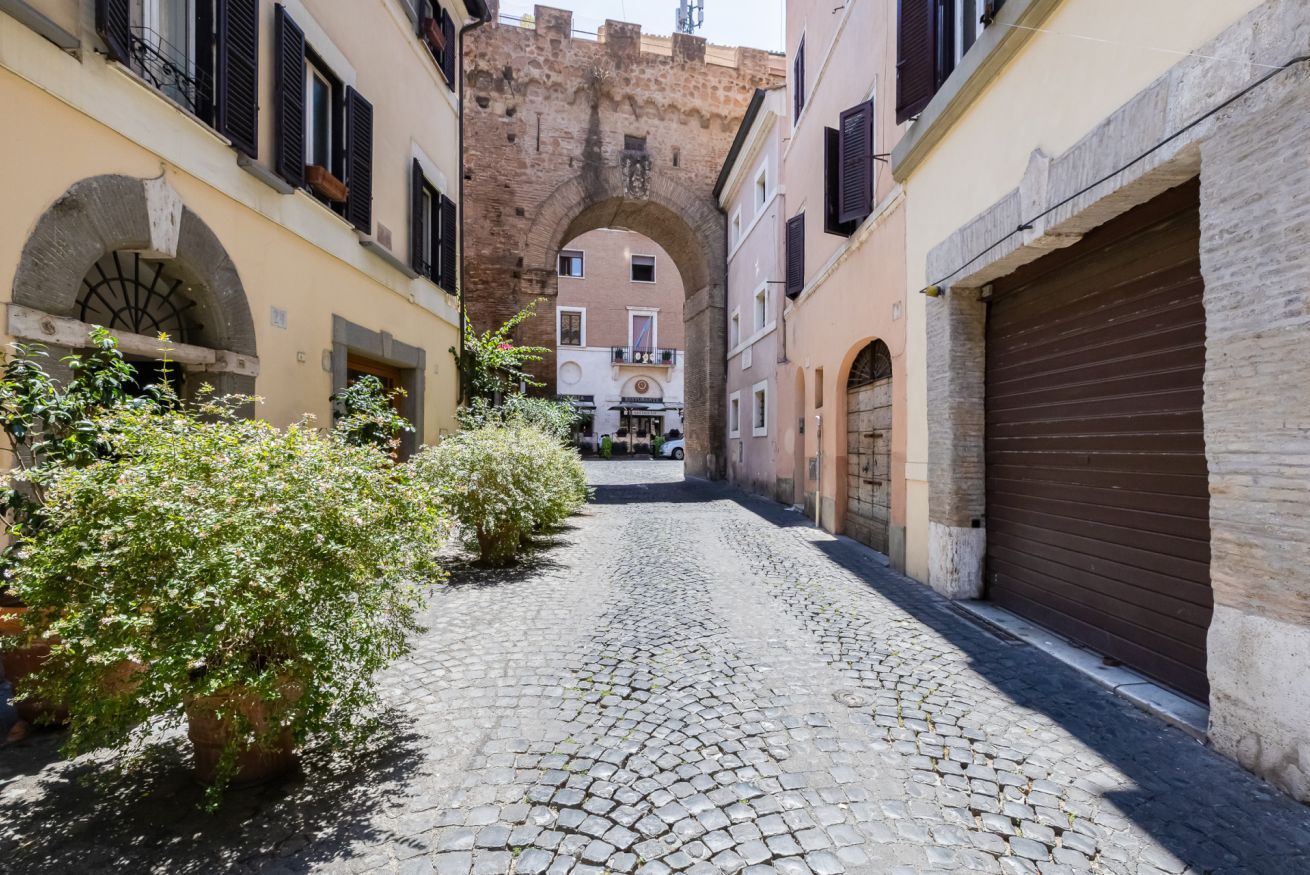 A cobblestone street with buildings on either side, an archway, and a small tree.
