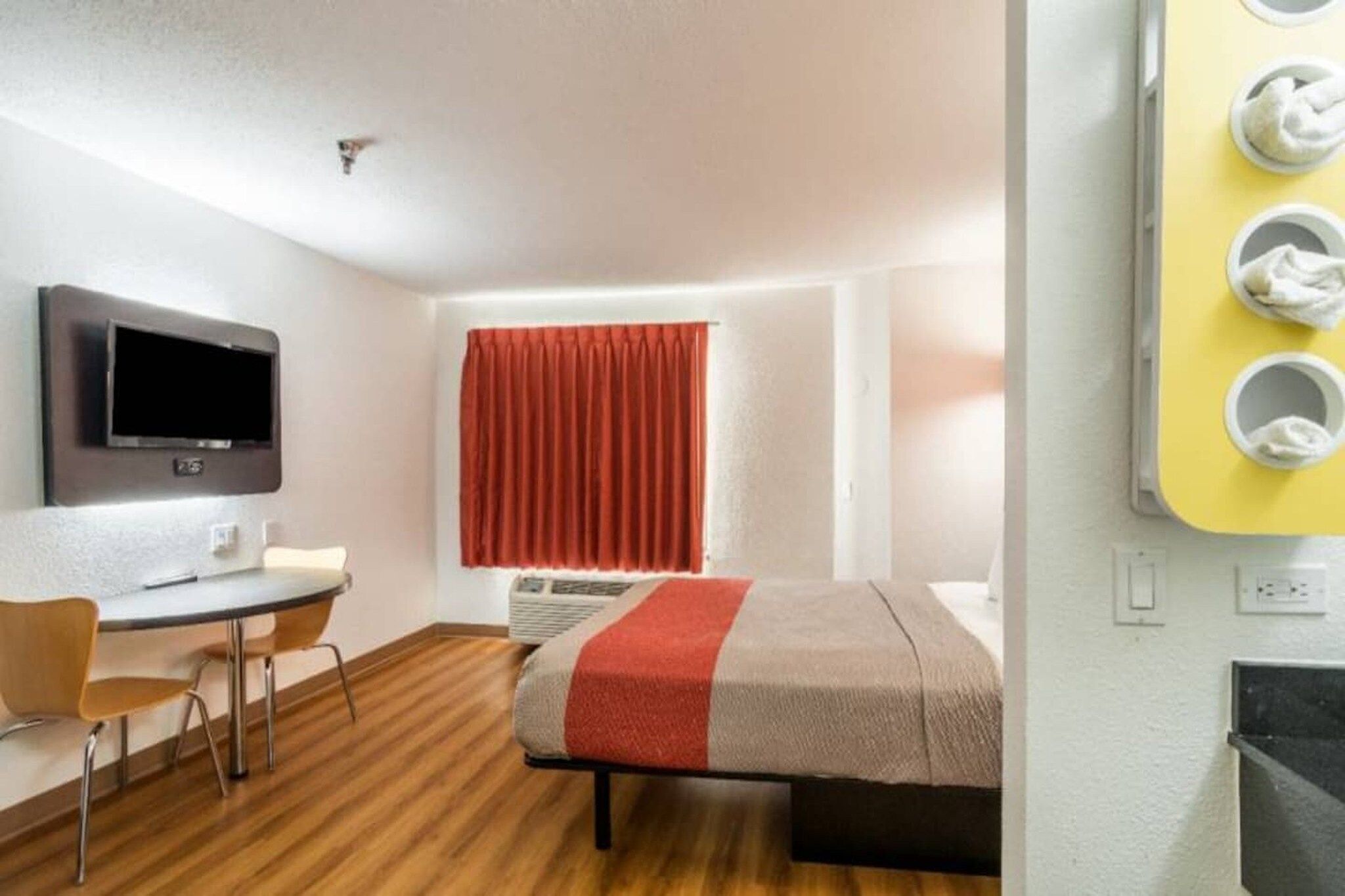 A hotel room with a bed, a desk, a chair, a television, a window with curtains, and a wall-mounted towel dispenser.