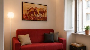 Un divano rosso con cuscini, una lampada da terra, un quadro, una finestra con vista e una piccola pianta sul davanzale.
