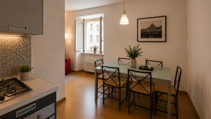 Una cucina moderna con piano cottura a gas, un'area pranzo con tavolo e sedie in vetro, un termosifone e una finestra con vista sugli edifici.