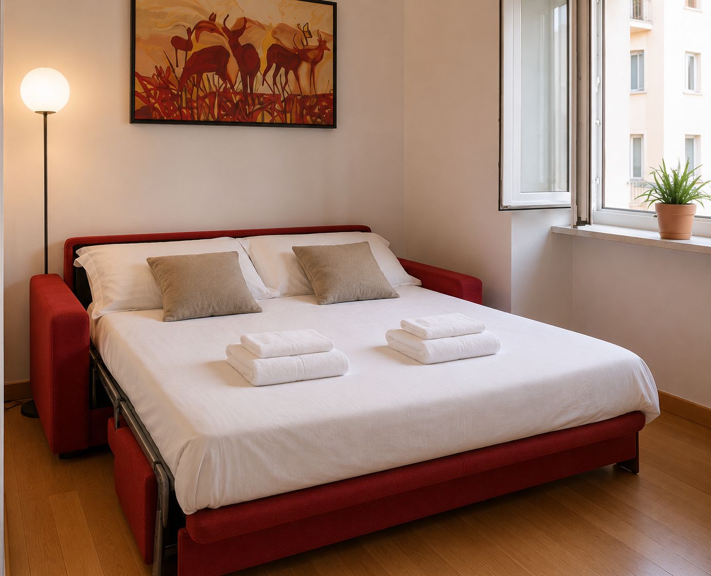 A hotel room with a bed, pillows, a bedside lamp, a painting, and a window with a plant.