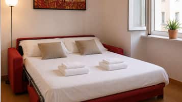 Habitación de hotel con cama, almohadas, lámpara de noche, un cuadro y ventana con planta.